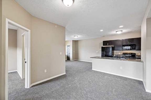Spacious apartment living area featuring neutral carpeting, soft beige walls, and an open view into the kitchen.