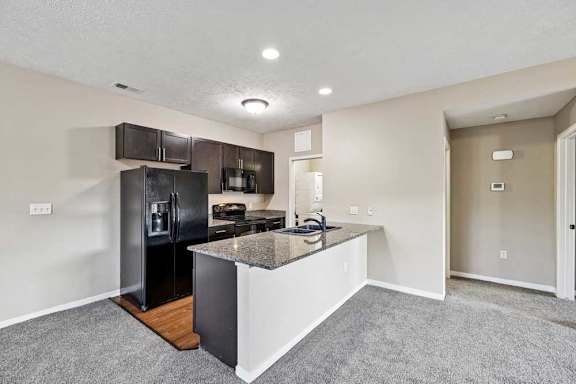 Open apartment kitchen featuring dark wood cabinets, black appliances, a granite island with sink, and carpeted living space.