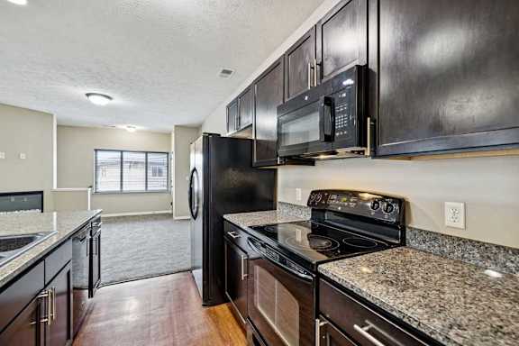 Modern apartment kitchen with granite countertops, dark cabinetry, black appliances, and an open view into the living area.