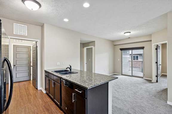 Open-concept apartment featuring a kitchen with dark wood cabinets and granite island, wood-style flooring, and a carpeted living area with sliding doors to a balcony.