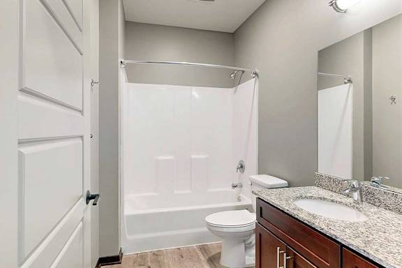 A bathroom with a granite vanity sink, and a white tub shower with a curved curtain rod.