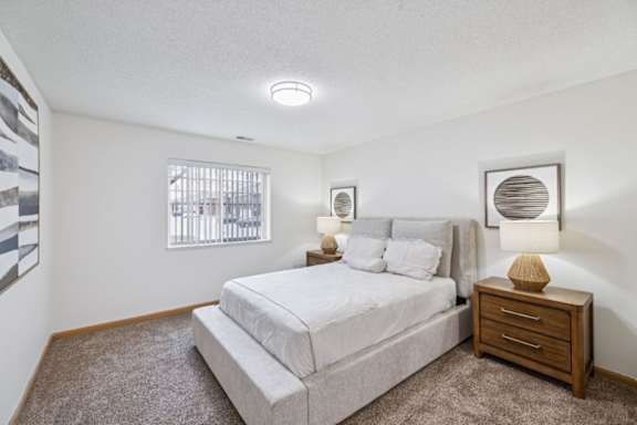 An apartment bedroom with a large bed, a nightstand, and a window with vertical blinds.