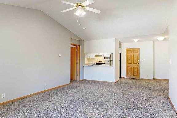 a carpeted living room with a vaulted ceiling and ceiling fan, next to the kitchen with a windowed countertop