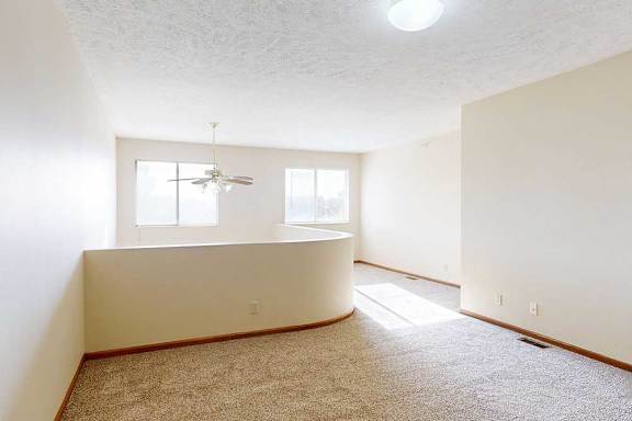 A carpeted loft bedroom with a view open to below, two bright windows, and a ceiling fan.