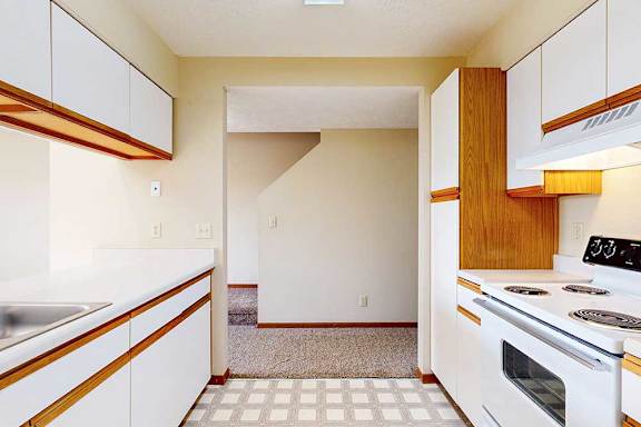 A galley kitchen with white cabinets with oak trim, white appliances, a double-basin sink, and a pass-through window countertop.