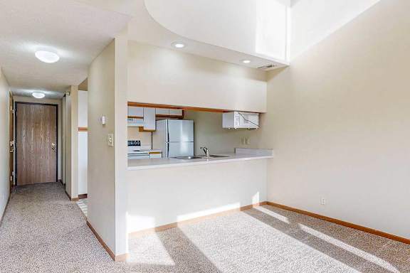 A carpeted living room with access to the kitchen via a pass-through window countertop, and a hallway connecting to the front entryway.