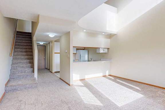 A carpeted living room with access to the kitchen via a pass-through window countertop, and a staircase to the upper loft area.