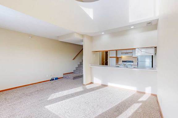 A carpeted living room with access to the kitchen via a pass-through window countertop, and a staircase to the upper loft area.