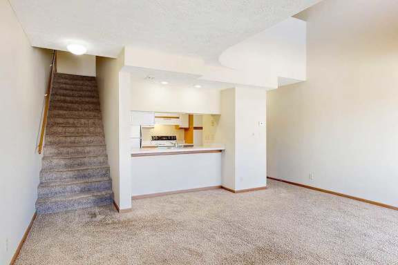 A carpeted living room with access to the kitchen via a pass-through window countertop, and a staircase to the upper loft area.
