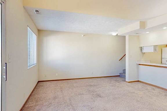 A carpeted living room with a window, access to the kitchen via a pass-through window countertop, and a staircase to the upper loft area.
