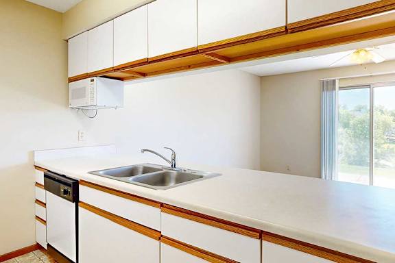 A galley kitchen featuring white cabinets with oak trim, a double basin sink, a mounted microwave, and a passthrough countertop window.