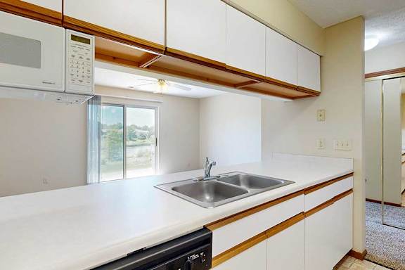 A galley kitchen featuring white cabinets with oak trim, a double basin sink, a mounted microwave, and a passthrough countertop window.