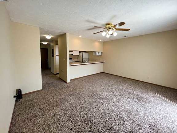 a living room with a ceiling fan and carpeted floor next to a window to the kitchen