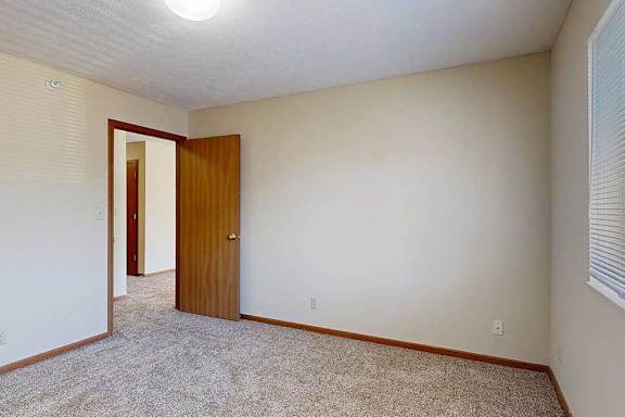 A bedroom with a carpeted floor, a window with blinds, and a wooden door.
