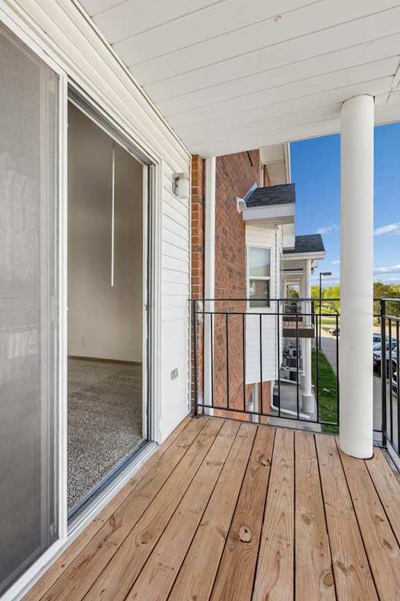 Covered apartment balcony with wood decking, a sliding glass door, brick accents, white support columns, and views of the surrounding apartment community.
