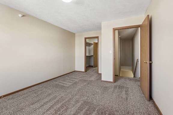Spacious apartment bedroom with neutral carpet, beige walls, wood-trimmed doors, and an open layout leading to the hallway and closet area.