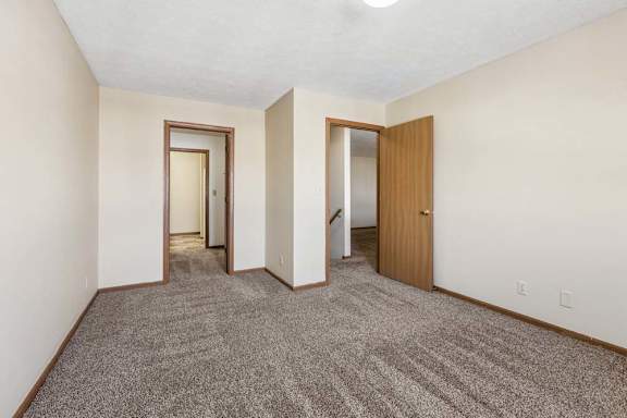 Spacious apartment bedroom with neutral carpet, beige walls, wood trim, and open doorways leading to the hallway, closet area, and staircase.