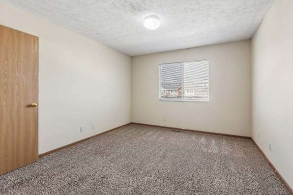 Bright, empty apartment bedroom with neutral carpet, beige walls, wood trim, and a window with blinds providing natural light.