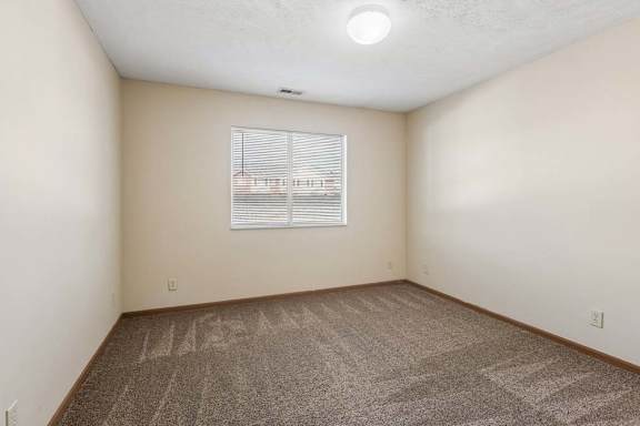 Bright, empty apartment bedroom with neutral carpet, beige walls, wood trim, and a window with blinds that brings in natural light.