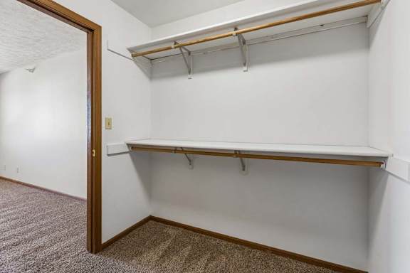 Walk-in apartment closet with carpeted flooring, dual hanging rods, upper shelving, and an open doorway connecting to the adjacent bedroom.