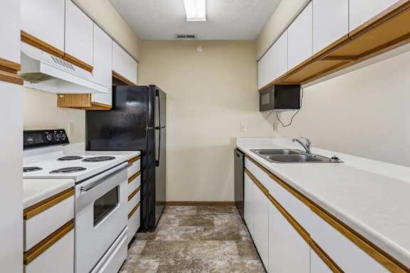 Galley-style apartment kitchen with white cabinets, white oven, black refrigerator, laminate countertops, a double-basin sink, and durable flooring in a functional layout.