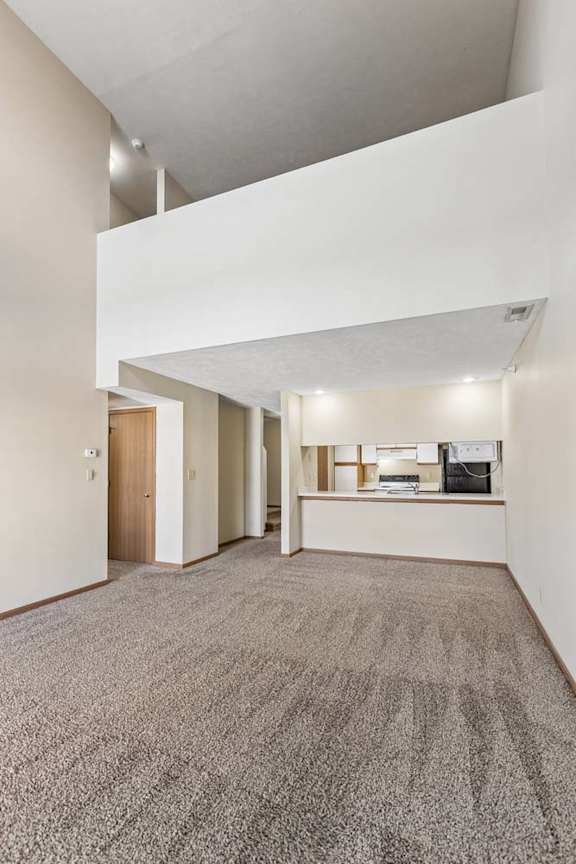 Spacious apartment living room with vaulted ceilings, neutral carpet, and an open-concept layout featuring a breakfast bar overlooking the kitchen.