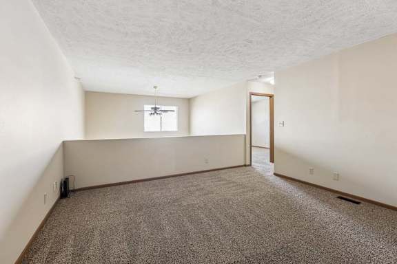 Spacious loft area in an apartment with neutral carpet, a half wall overlooking the lower level, a window for natural light, and an open doorway leading to the hallway.