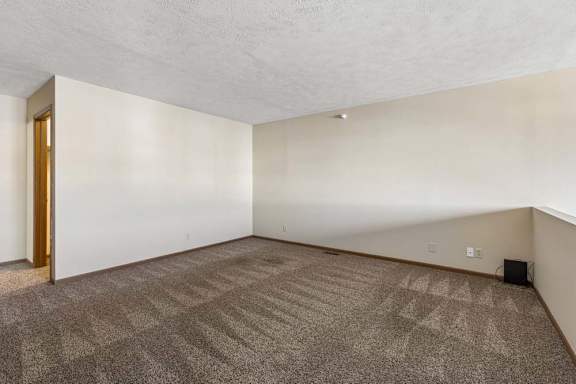 Spacious apartment loft with neutral carpet, a half wall overlooking the living area below, and an open layout with access to the adjoining hallway.