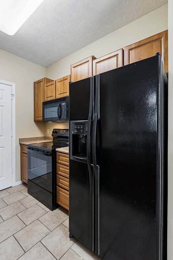 A black refrigerator stands in a kitchen with wooden cabinets and a microwave.
