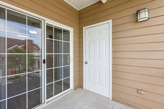 Covered apartment balcony with sliding glass doors, tan siding, white entry door, and outdoor wall light.