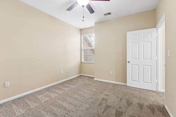 Bright, empty apartment bedroom with beige walls, carpet, ceiling fan, and window with blinds.