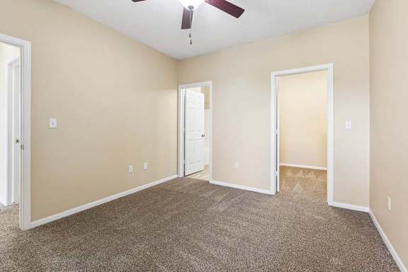 Carpeted apartment bedroom with beige walls, ceiling fan, and open doorways to nearby rooms.