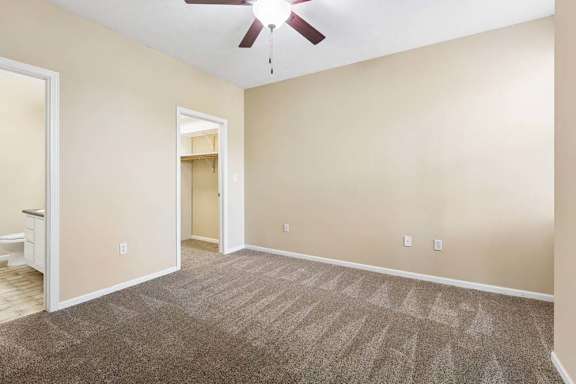 Empty apartment bedroom with plush carpet, ceiling fan, and open access to bathroom and walk-in closet.