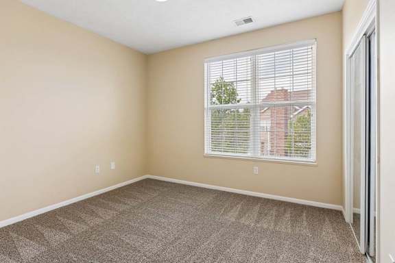 Bright apartment bedroom with large window, beige walls, carpet, and mirrored closet doors.