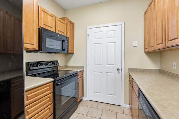 Galley-style apartment kitchen with wood cabinets, black appliances, oven, microwave, and tile floor.