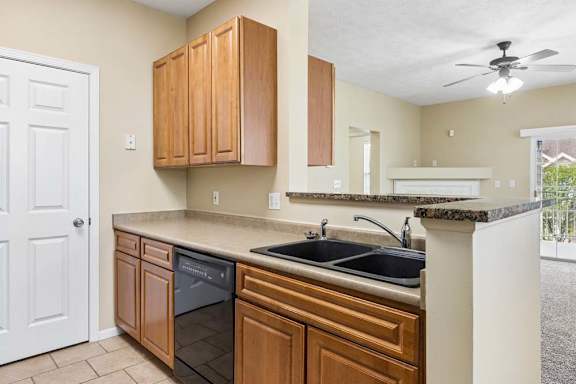 Apartment kitchen with wood cabinetry, double sink, black dishwasher, breakfast bar, and view into living room.