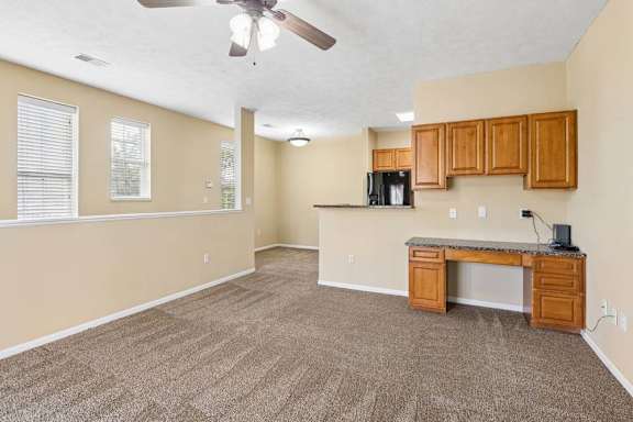 Open apartment living area with carpet, built-in desk cabinetry, wood cabinets, and windows with blinds.