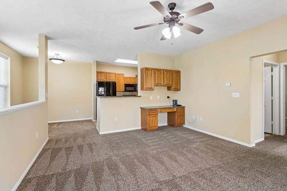 Spacious apartment living room with ceiling fan, carpet, built-in desk, and open kitchen with wood cabinets.