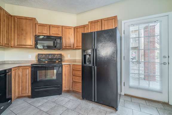 A black refrigerator stands in a kitchen with wooden cabinets and a black microwave.