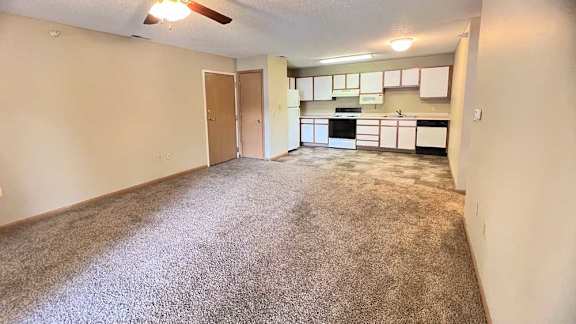 a spacious living room with a carpeted floor and a ceiling fan, and a kitchen with white and brown cabinets in the background
