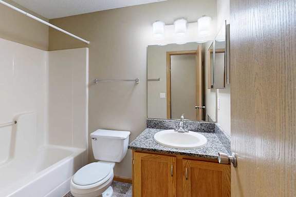 Bathroom interior with white tub and shower surround, single-sink vanity with speckled countertop, wood cabinets, and large mirror with overhead lighting.