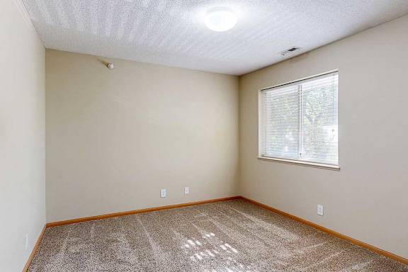 Bright bedroom with beige walls, plush carpet, wood trim, and a large window with blinds bringing in natural light.