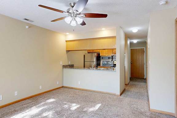 Open-concept living and dining area with ceiling fan, plush carpet, breakfast bar, and kitchen with wood cabinetry and stainless-steel appliances.