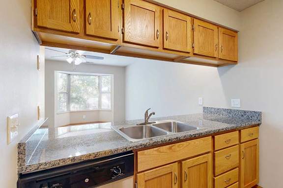 Open kitchen counter with double sink, wood cabinets, and breakfast bar connecting to a bright living area with bay window.