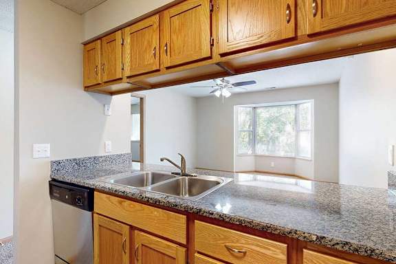 Open kitchen counter with double sink, wood cabinets, and breakfast bar connecting to a bright living area with bay window.