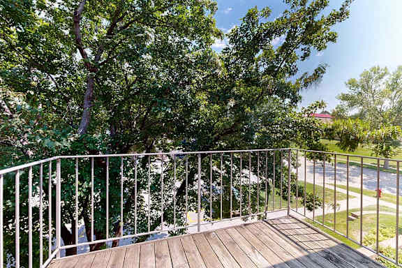 Private apartment balcony with wood decking and metal railing overlooking mature trees and a residential street.