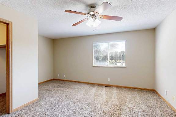 Spacious bedroom with plush carpet, neutral walls, ceiling fan with light, and wide window with blinds.