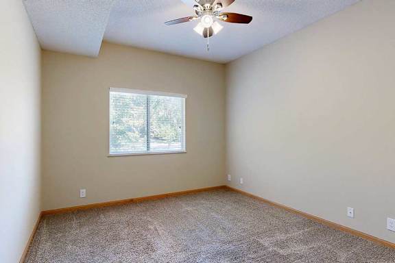 Empty bedroom with wide window and blinds, soft carpet flooring, and ceiling fan for added comfort.