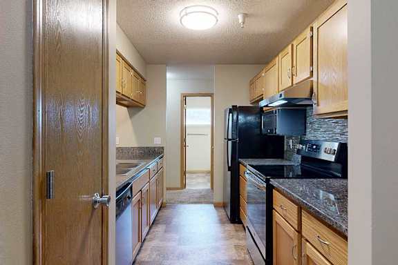 Galley-style kitchen with wood cabinets, granite-style countertops, black appliances, tile backsplash, and vinyl flooring.