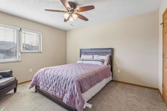 Bright apartment bedroom with neutral carpet, large corner windows, ceiling fan, and a queen-size bed, offering a comfortable and airy interior.